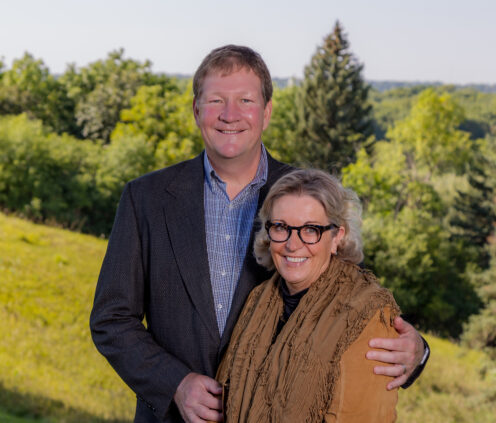Outdoor photo of Brian and Denise Aamlid, smiling next to one another while Brian has his arm around Denise.
