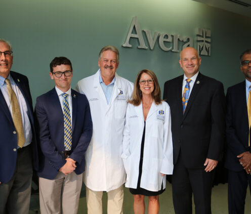 Photo of Kevin and Lorie Haarberg smiling, wearing pharmacy whites that were given to them as gifts from the College. Also photographed with them is President Dunn, Dean Dan Hansen, Provost Dennis Hedge, and other member of the college.
