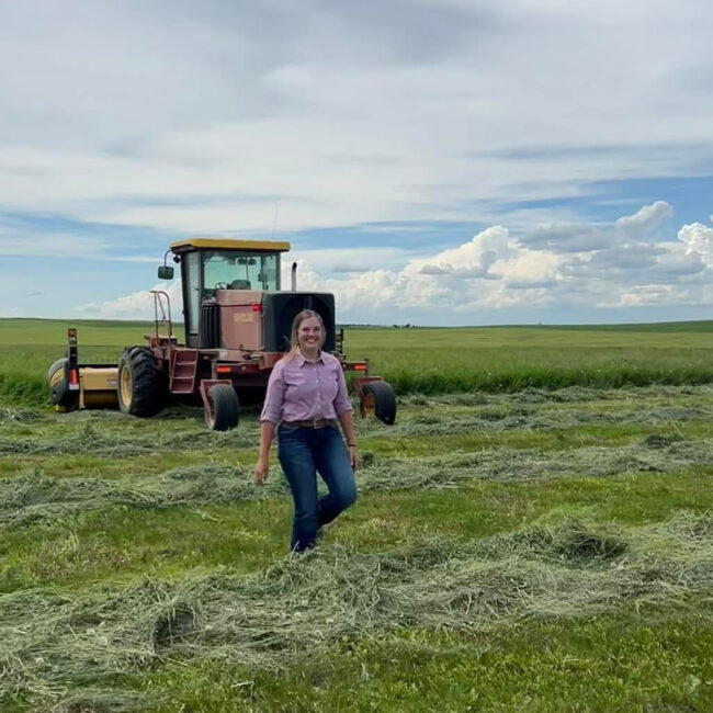 Abby stands in a field with a beautiful blue sky and a tractor behind her.