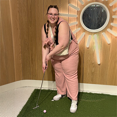 Allison Fesler smiles while holding a golf club, putting.