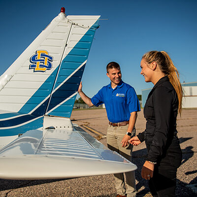 Two SDSU aviation students stand near the tail of an airplane, adorned with blue strips and the SDSU logo.
