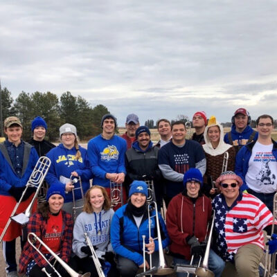 Large group of students holding trombones, dressed in patriot costumes/clothes during a rehearsal.
