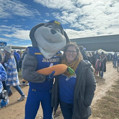 Jennifer Benson, at football tailgating, smiles next to Jack the Jackrabbit who is holding his stuffed carrot.