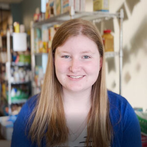 Headshot of Keri Pappas sitting in Jack's Cupboard, with shelves of food in the background.