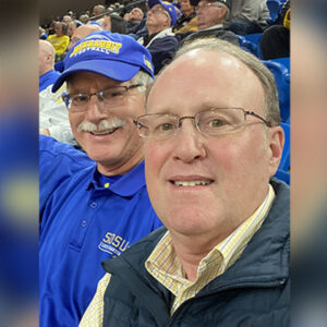 Craig Carson and Dave Schaefer smile at the camera while attending a Jackrabbit basketball game.