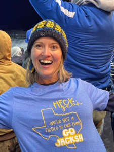 Julie Mooney, smiling ear to ear, as she reveals her shirt that says "Heck yeah, we've got a fiddle in our band. Go Jacks!"