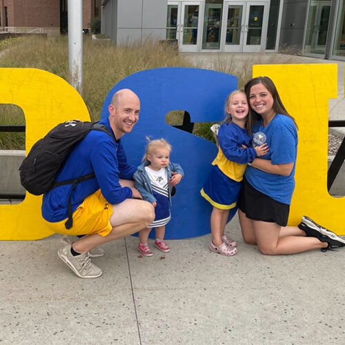 Allison and her husband smiling infront of large SDSU wooden letters, smiling with their two daughters.