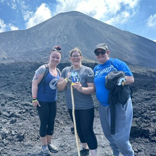 Jennifer Benson stands with two friends while on a hike with a large mountain and blue, cloudy sky in the background.