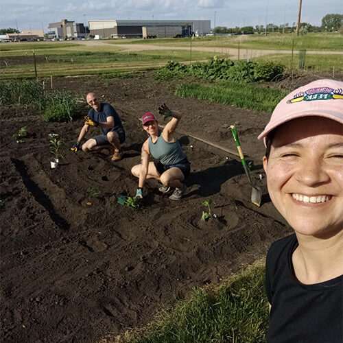 Anne Karabon waving at the camera while planting a garden at the Brookings community gardens with her partner, Matt, and GA, Laura.