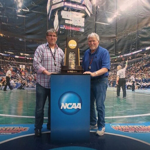 Frank and his son, Steve, smiling for the camera at a NCAA wrestling tournament, posing with a large trophy.