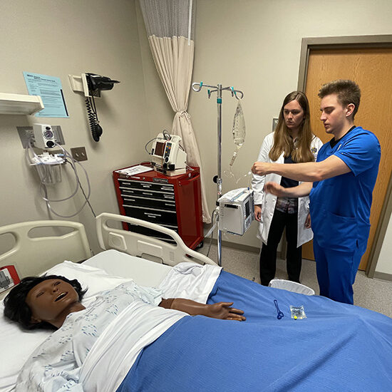 Male SDSU nursing student working with a professor while overlooking a mannequin patient.