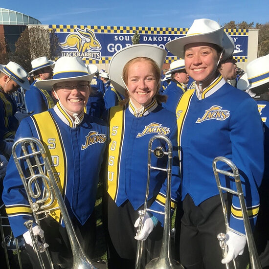 Jessica smiles while standing with two friends, all holding trombones in their Pride uniforms.