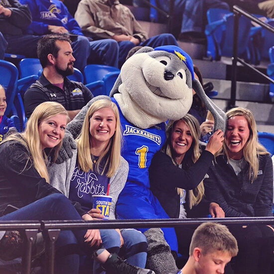 Cassie and three other females smiling at a basketball game, while Jack the Jackrabbit sits with them, holding his floppy ear over Cassie.