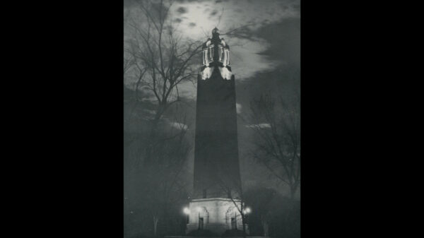 Black and white photo of the Campanile at night, with a cloudy sky and moon shining down over the Campanile.