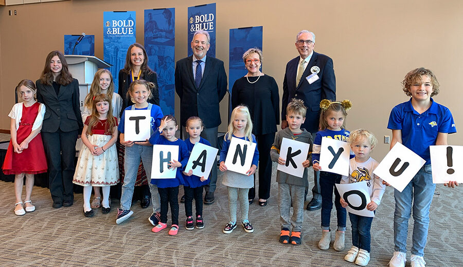 Tate Profilet and Mary DeJong pose with Dr. Carie Green, her children, and President Dunn during the investiture ceremony. Eight children stand in front of them, holding up letters spelling out "thank you!"