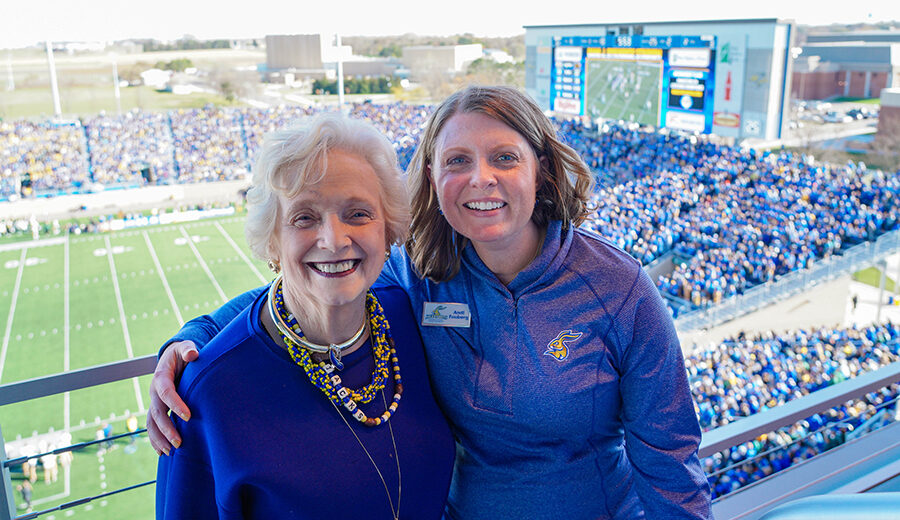Peggy Gordon Miller and Andi Fouberg smiling for the camera at a SDSU football game, all dressed in blue and Jackrabbit gear.