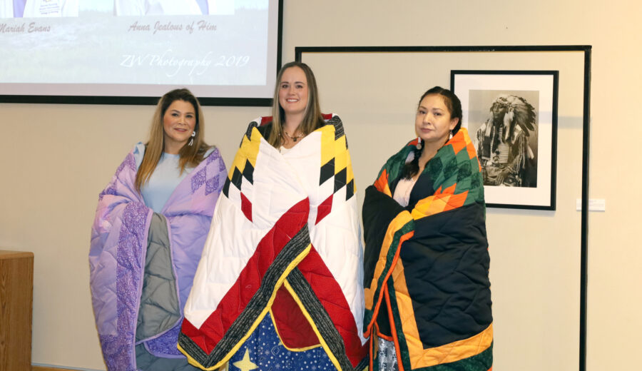 3 nursing students wrapped in star quilts at their honoring ceremony. Anna Jealous of Him is pictured far right.