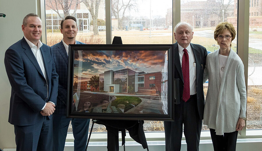 Larry and Diane Ness stand with their two sons next to a large framed photograph of the Ness School.