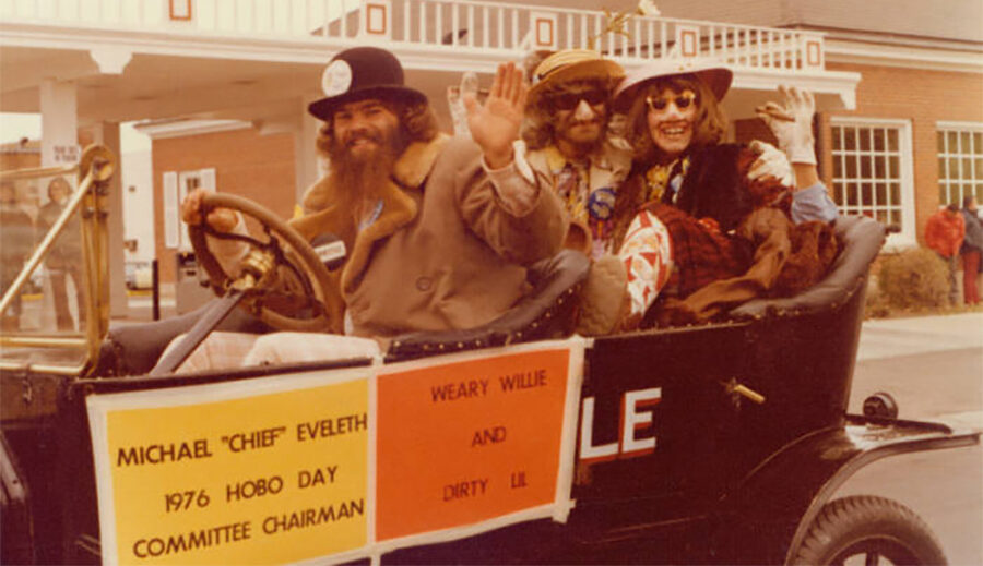 The first Dirty Lil and Students' Association president, Barb Strandell, and Weary Wil, Jack Soukup, and riding in the Bummobile which is being driven by Michael 'Chief' Eveleth, 1976 Hobo Day committee chairman and Grand Pooba