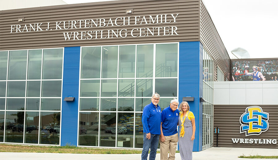 Frank Kurtenbach and his daughter and son smiling, standing side by side outside and infront of the Frank J. Kurtenbach Family Wrestling Center